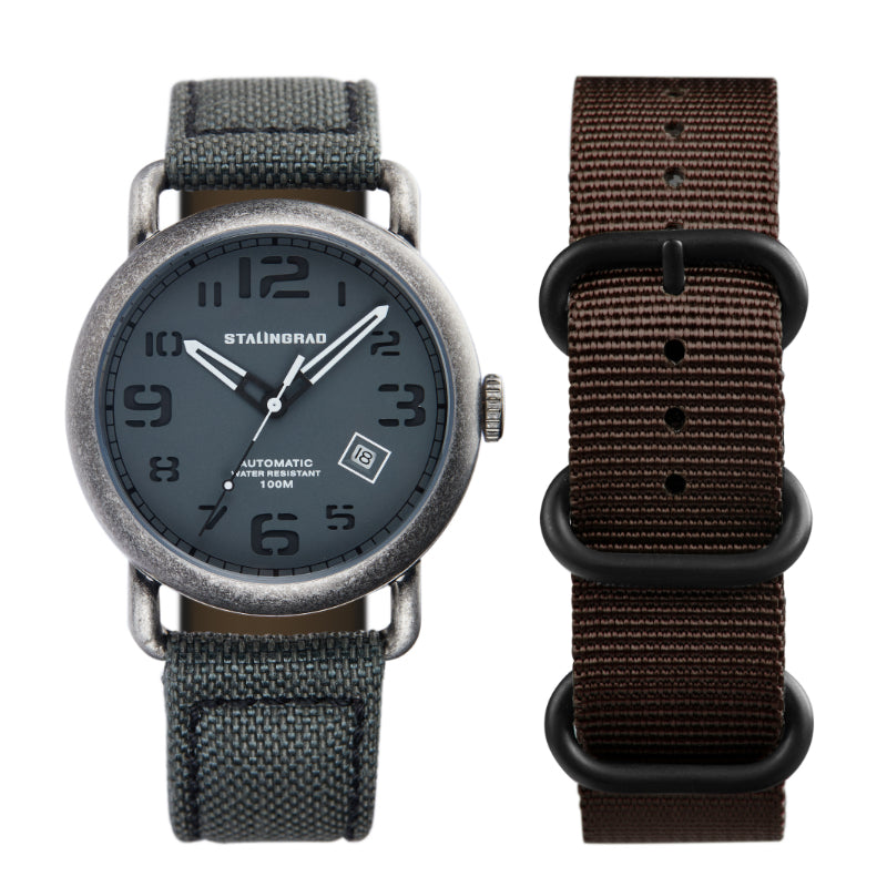 Stalingrad Rodim Watch with a sandwich dial. Grey dial with white hands, Silver case and a grey cordura strap with a brown nato strap next to it front view of watch on white background.