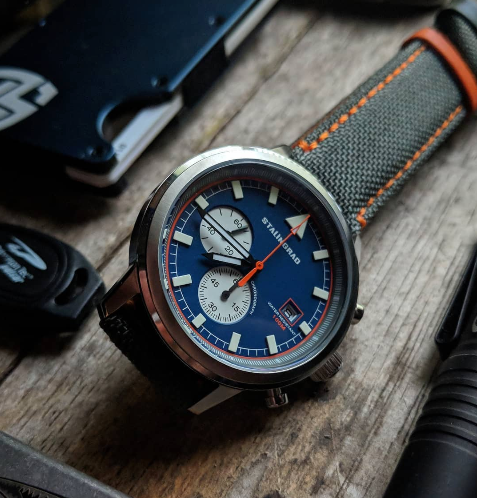 Stalingrad trooper chronograph watch with silver case, blue dial and grey Cordura straps with orange stitching, front view, on a wooden table 